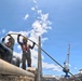 Two workers in safety gear are handling a heavy rope or mooring line at what appears to be a maritime or shipyard facility. They are positioned near large mooring bollards on a dock or vessel, with industrial cranes visible in the background. The scene suggests active docking, mooring, or rigging operations at a port or naval facility.
