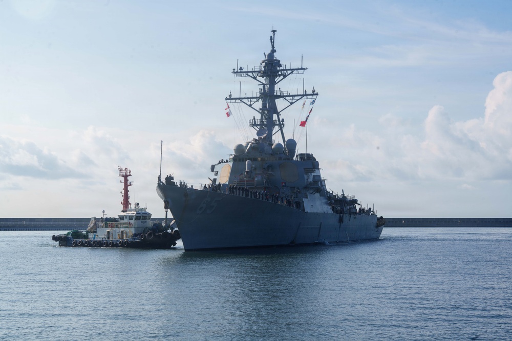 A large U.S. Navy Arleigh Burke-class guided-missile destroyer is maneuvering in a harbor or port, assisted by a tugboat on its port side. The warship is painted in standard naval gray and has personnel visible along the deck rails. Multiple flags and radar/communications arrays are visible on the mast superstructure.