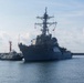 A large U.S. Navy Arleigh Burke-class guided-missile destroyer is maneuvering in a harbor or port, assisted by a tugboat on its port side. The warship is painted in standard naval gray and has personnel visible along the deck rails. Multiple flags and radar/communications arrays are visible on the mast superstructure.