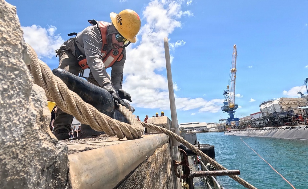 A worker in full safety gear is handling heavy mooring lines on a concrete pier or dock structure at a naval shipyard. In the background, a submarine is visible docked alongside industrial cranes and shipyard facilities. The scene appears to depict a ship mooring or line-handling operation in a military or industrial port environment.