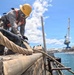 A worker in full safety gear is handling heavy mooring lines on a concrete pier or dock structure at a naval shipyard. In the background, a submarine is visible docked alongside industrial cranes and shipyard facilities. The scene appears to depict a ship mooring or line-handling operation in a military or industrial port environment.