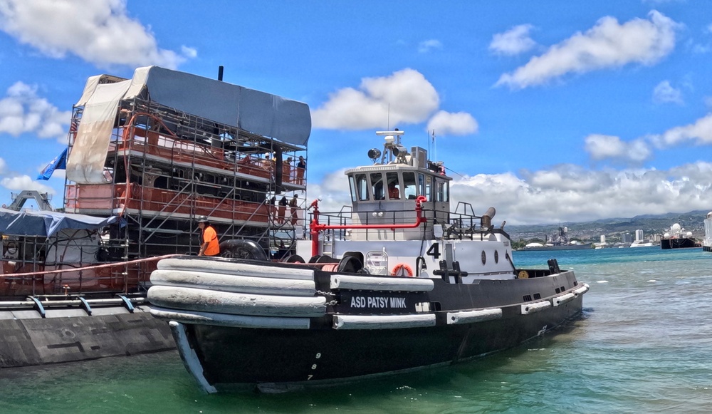 A tugboat named 'ASD Patsy Mink' (numbered 4) is positioned alongside a large barge or vessel undergoing restoration or construction work, with scaffolding and tarpaulin coverings visible. The scene appears to be in a harbor, likely Pearl Harbor, Hawaii, based on the recognizable skyline and turquoise water in the background. Workers in safety gear are visible on the scaffolding structure.