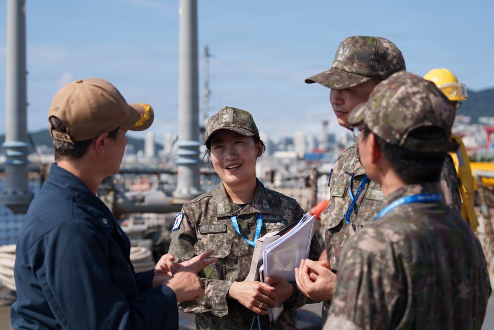 Four individuals are engaged in a discussion on what appears to be a naval vessel or port facility deck. Three individuals are wearing camouflage military uniforms consistent with Republic of Korea (ROK) Armed Forces, while one person on the left wears a navy blue uniform with a tan baseball cap. The central figure, a female ROK military officer, is holding documents and appears to be leading the conversation.