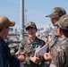 Four individuals are engaged in a discussion on what appears to be a naval vessel or port facility deck. Three individuals are wearing camouflage military uniforms consistent with Republic of Korea (ROK) Armed Forces, while one person on the left wears a navy blue uniform with a tan baseball cap. The central figure, a female ROK military officer, is holding documents and appears to be leading the conversation.