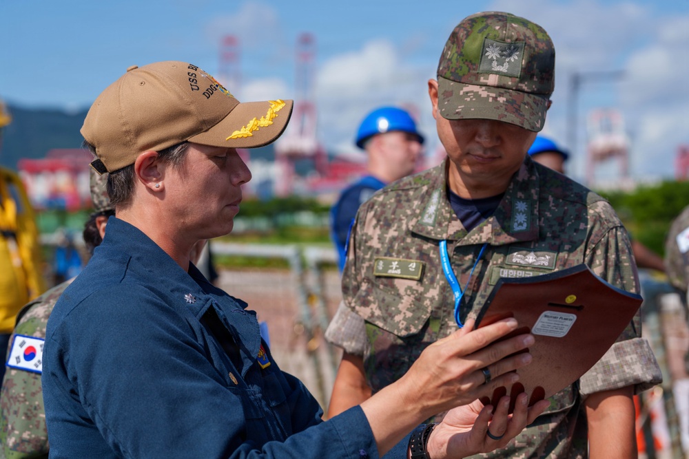 Two military personnel — one appearing to be a U.S. Navy sailor wearing a tan cap with gold insignia and a South Korean flag patch, and one in Republic of Korea (ROK) Army digital camouflage uniform — are reviewing a document or tablet together. The interaction appears to be a joint military coordination or briefing activity, likely at a port or industrial facility. The setting and uniforms suggest a bilateral military exercise or port visit.