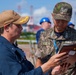Two military personnel — one appearing to be a U.S. Navy sailor wearing a tan cap with gold insignia and a South Korean flag patch, and one in Republic of Korea (ROK) Army digital camouflage uniform — are reviewing a document or tablet together. The interaction appears to be a joint military coordination or briefing activity, likely at a port or industrial facility. The setting and uniforms suggest a bilateral military exercise or port visit.