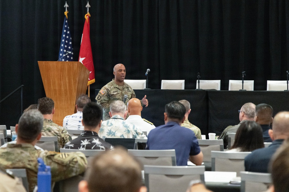 A U.S. Army officer in camouflage uniform is addressing an audience at a formal military briefing or conference. The speaker stands near a wooden podium flanked by the U.S. flag and a red Army general officer flag with stars. The audience consists of a mix of military personnel in uniform and civilians seated in rows.