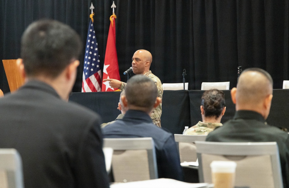 A military officer in camouflage uniform is speaking into a microphone at what appears to be a formal briefing or conference setting. He is gesturing with his finger toward the audience, which consists of both military personnel and civilians seated at tables. Two flags — the United States flag and a red U.S. Army flag — are visible in the background.