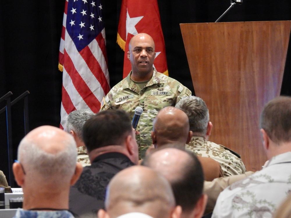 A U.S. Army officer with the name tape 'CLARK' and rank insignia visible on his uniform is addressing an audience at a formal military briefing or conference. He is holding a microphone and standing in front of a wooden podium, flanked by the U.S. flag and a red Army general officer flag. The audience consists of multiple military personnel in various uniforms seated with their backs to the camera.