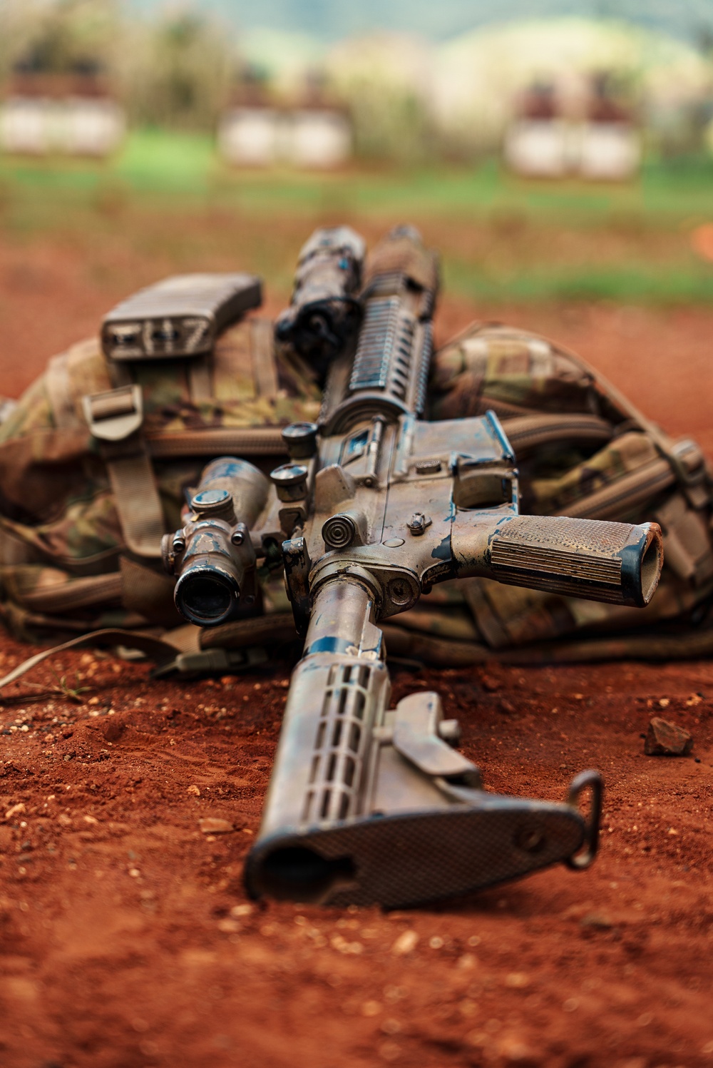 A heavily worn assault rifle, consistent with an M4/AR-15 platform, is laid on red laterite soil atop a camouflage tactical vest or plate carrier. The weapon shows significant wear and blue paint markings, possibly indicating use in training exercises. The background is blurred but suggests an outdoor military or training environment with structures and vegetation visible.