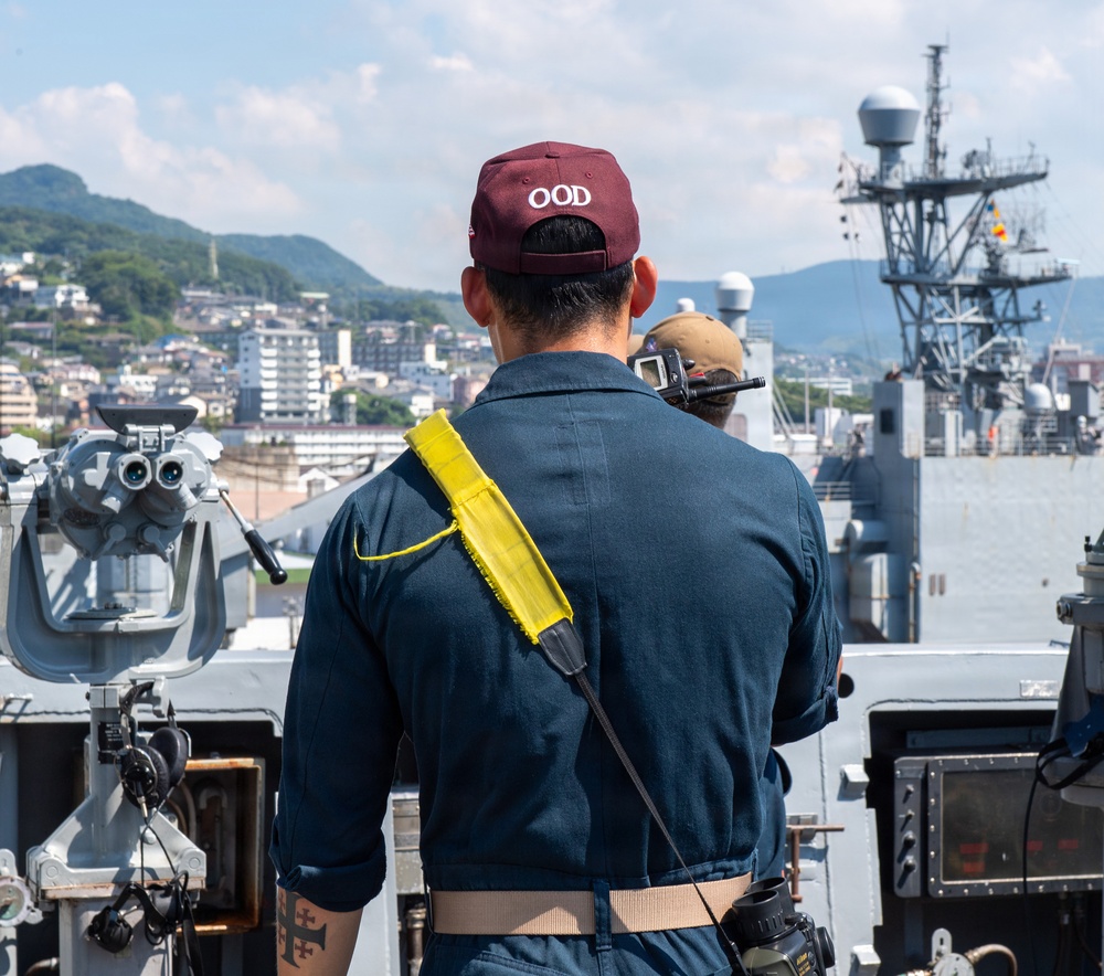 A naval officer wearing a maroon cap labeled 'OOD' (Officer of the Deck) stands on the bridge wing of a warship, facing forward toward a harbor. The officer is equipped with a yellow signal/communication satchel and a radio, overseeing ship operations. A coastal city with green hills is visible in the background, along with another naval vessel moored nearby.
