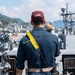 A naval officer wearing a maroon cap labeled 'OOD' (Officer of the Deck) stands on the bridge wing of a warship, facing forward toward a harbor. The officer is equipped with a yellow signal/communication satchel and a radio, overseeing ship operations. A coastal city with green hills is visible in the background, along with another naval vessel moored nearby.