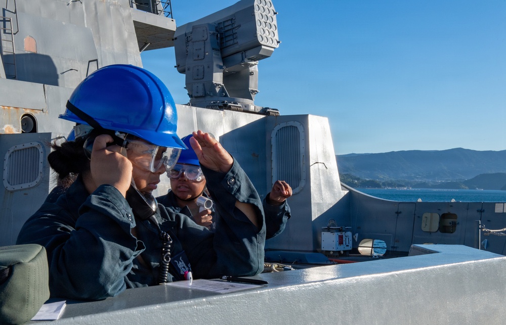 Two naval personnel wearing blue hard hats and safety goggles are stationed on the deck of a warship, with one using a radio/communication handset. Behind them is a visible weapons system, likely a Phalanx Close-In Weapon System (CIWS) or similar naval defense system. The scene appears to be during a port operation or underway evolution with mountainous coastline visible in the background.