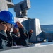 Two naval personnel wearing blue hard hats and safety goggles are stationed on the deck of a warship, with one using a radio/communication handset. Behind them is a visible weapons system, likely a Phalanx Close-In Weapon System (CIWS) or similar naval defense system. The scene appears to be during a port operation or underway evolution with mountainous coastline visible in the background.