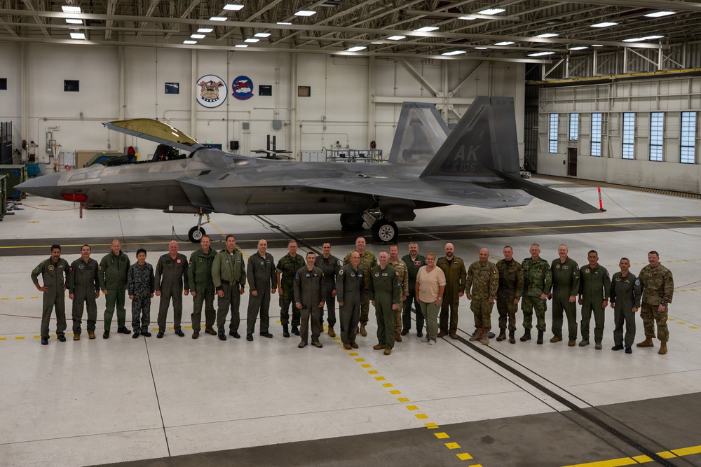 A group of approximately 25 military personnel from multiple nations pose for a formal group photograph inside a large aircraft hangar in front of an F-22 Raptor stealth fighter jet. The aircraft bears the tail code 'AK' indicating assignment to Alaska (likely Elmendorf-Richardson Air Force Base). Personnel are wearing various military flight suits and uniforms representing different countries and service branches.