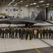 A group of approximately 25 military personnel from multiple nations pose for a formal group photograph inside a large aircraft hangar in front of an F-22 Raptor stealth fighter jet. The aircraft bears the tail code 'AK' indicating assignment to Alaska (likely Elmendorf-Richardson Air Force Base). Personnel are wearing various military flight suits and uniforms representing different countries and service branches.