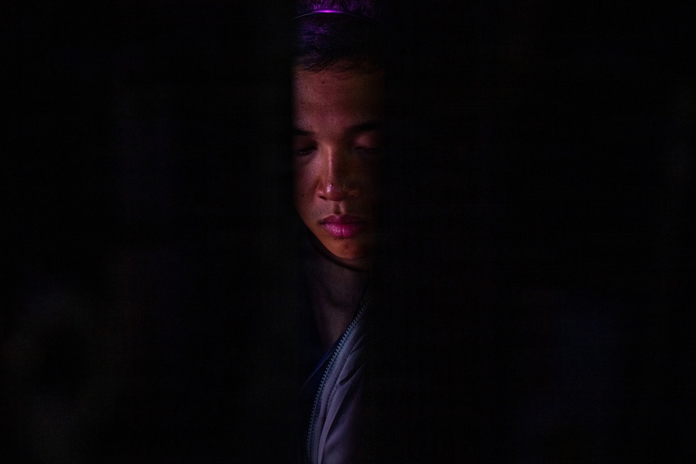 A dramatic low-key portrait of a young person partially illuminated against an almost entirely dark background. The face is lit from above and slightly to the right, revealing facial features with eyes appearing closed or downcast. A faint purple/magenta light is visible at the top of the frame, and the subject appears to be wearing a zip-up jacket.