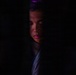 A dramatic low-key portrait of a young person partially illuminated against an almost entirely dark background. The face is lit from above and slightly to the right, revealing facial features with eyes appearing closed or downcast. A faint purple/magenta light is visible at the top of the frame, and the subject appears to be wearing a zip-up jacket.