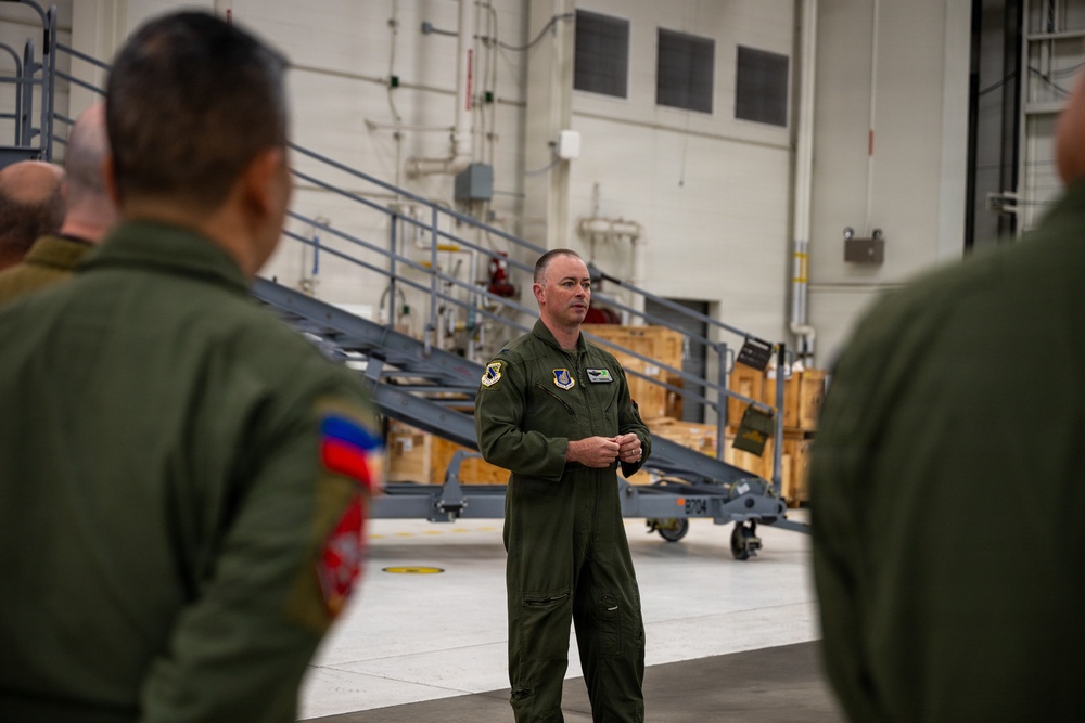 A military briefing or orientation is taking place inside what appears to be an aircraft hangar or maintenance facility. A central figure in olive green flight suit with US Air Force patches is addressing a small group of similarly uniformed personnel. The setting includes industrial equipment, staircases, and storage crates typical of a military aviation maintenance environment.