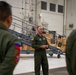 A military briefing or orientation is taking place inside what appears to be an aircraft hangar or maintenance facility. A central figure in olive green flight suit with US Air Force patches is addressing a small group of similarly uniformed personnel. The setting includes industrial equipment, staircases, and storage crates typical of a military aviation maintenance environment.