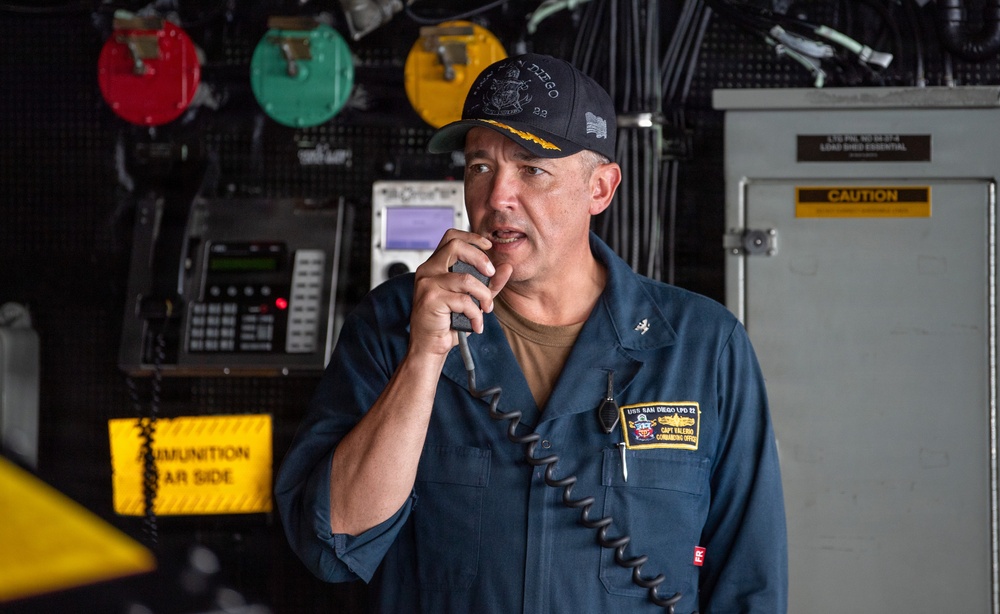 A U.S. Navy officer, identified by his name tag as 'CAPT VALERO, COMMANDING OFFICER' aboard the USS San Diego LPD 22, is speaking into a shipboard intercom or sound-powered phone handset. He is wearing a dark blue Navy coverall uniform and a black ball cap with gold insignia. The setting appears to be a shipboard compartment or hangar bay area with various equipment and warning signs visible in the background.