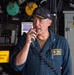 A U.S. Navy officer, identified by his name tag as 'CAPT VALERO, COMMANDING OFFICER' aboard the USS San Diego LPD 22, is speaking into a shipboard intercom or sound-powered phone handset. He is wearing a dark blue Navy coverall uniform and a black ball cap with gold insignia. The setting appears to be a shipboard compartment or hangar bay area with various equipment and warning signs visible in the background.