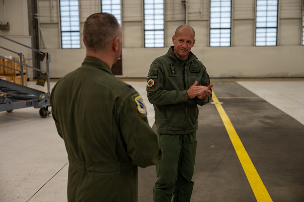 Two military personnel in olive green flight suits are engaged in conversation inside a large aircraft hangar. Both individuals wear unit patches on their sleeves, suggesting they are aviators or aircrew members. The hangar interior is well-lit with large windows and features a yellow safety line painted on the floor.