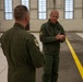 Two military personnel in olive green flight suits are engaged in conversation inside a large aircraft hangar. Both individuals wear unit patches on their sleeves, suggesting they are aviators or aircrew members. The hangar interior is well-lit with large windows and features a yellow safety line painted on the floor.