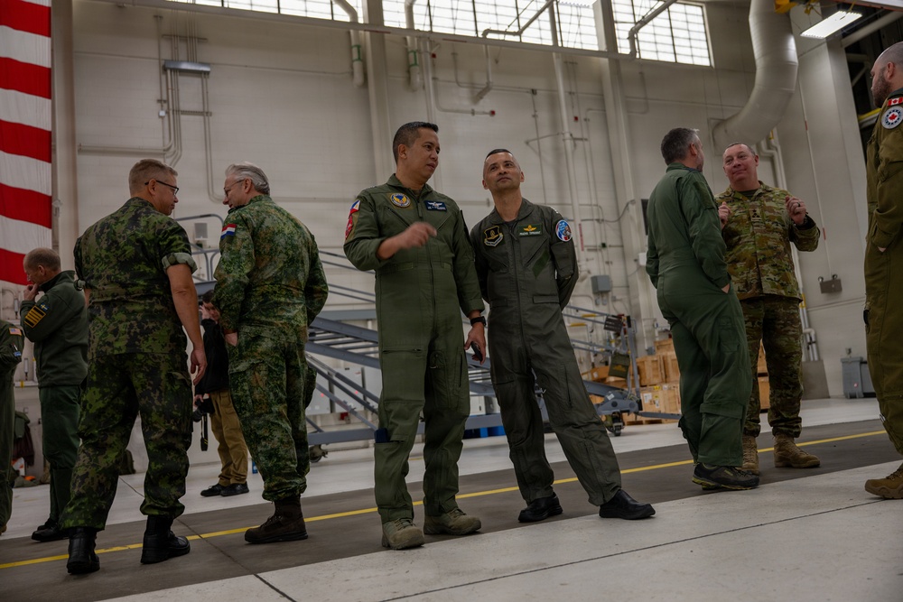 A group of military personnel from multiple nations are gathered inside a large aircraft hangar, engaged in informal conversations. The individuals are wearing various military uniforms including flight suits and camouflage patterns, suggesting a multinational military event or briefing. An American flag is partially visible on the left wall of the hangar.
