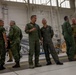 A group of military personnel from multiple nations are gathered inside a large aircraft hangar, engaged in informal conversations. The individuals are wearing various military uniforms including flight suits and camouflage patterns, suggesting a multinational military event or briefing. An American flag is partially visible on the left wall of the hangar.