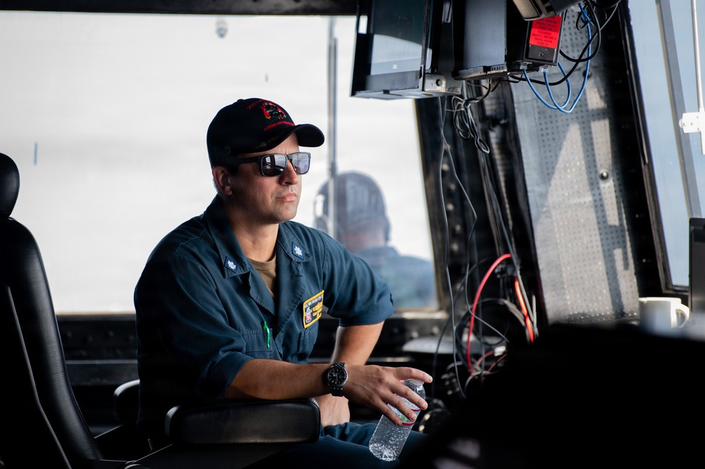 A military or naval officer in blue utility coveralls sits in what appears to be a vessel's bridge or command station. He is wearing a black baseball cap with insignia, sunglasses, and a watch, while holding a water bottle. The setting contains multiple monitors, wiring, and control equipment consistent with a naval or coast guard vessel.