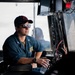 A military or naval officer in blue utility coveralls sits in what appears to be a vessel's bridge or command station. He is wearing a black baseball cap with insignia, sunglasses, and a watch, while holding a water bottle. The setting contains multiple monitors, wiring, and control equipment consistent with a naval or coast guard vessel.