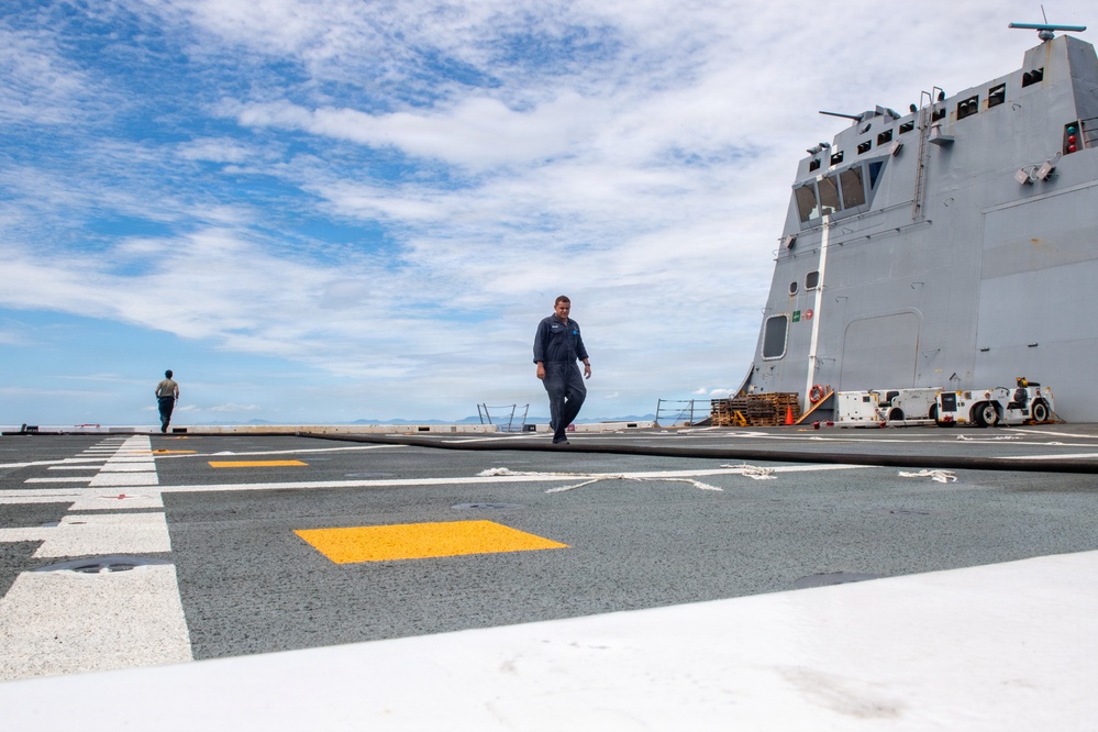 The image shows the flight deck of a U.S. Navy Littoral Combat Ship (LCS), identifiable by its distinctive angular superstructure. Two personnel in dark coveralls are walking across the deck, which features standard flight deck markings including white lines and a yellow rectangle. Various deck equipment including orange traffic cones, wooden pallets, and a deck tractor are visible near the superstructure.