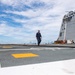 The image shows the flight deck of a U.S. Navy Littoral Combat Ship (LCS), identifiable by its distinctive angular superstructure. Two personnel in dark coveralls are walking across the deck, which features standard flight deck markings including white lines and a yellow rectangle. Various deck equipment including orange traffic cones, wooden pallets, and a deck tractor are visible near the superstructure.