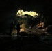 Two silhouetted figures stand inside a dark cave, facing toward a natural opening or skylight that allows bright natural light to filter in from above. The illuminated opening reveals green vegetation and rocky formations at the cave's upper edge. The contrast between the dark cave interior and the bright opening creates a dramatic chiaroscuro effect.