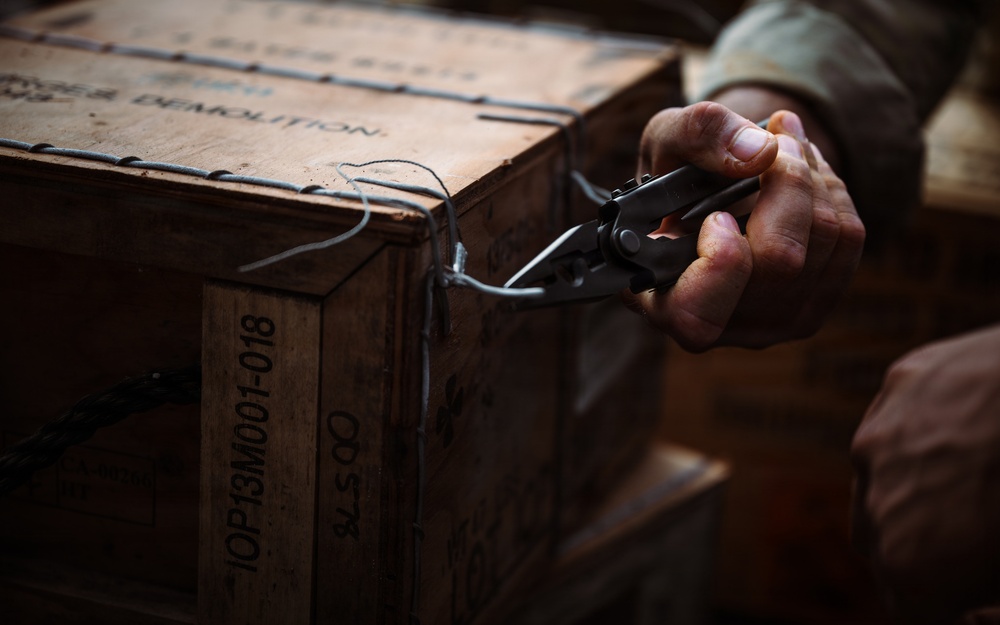 A person in military or tactical clothing is using wire cutters or pliers on a wooden crate stenciled with 'DEMOLITION' and a lot number 'IOP13M001-018'. The crate appears to be a military-style ammunition or demolition supply box secured with wire banding. The image is a close-up, dark-toned photograph focusing on the hands and the crate.