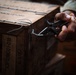 A person in military or tactical clothing is using wire cutters or pliers on a wooden crate stenciled with 'DEMOLITION' and a lot number 'IOP13M001-018'. The crate appears to be a military-style ammunition or demolition supply box secured with wire banding. The image is a close-up, dark-toned photograph focusing on the hands and the crate.
