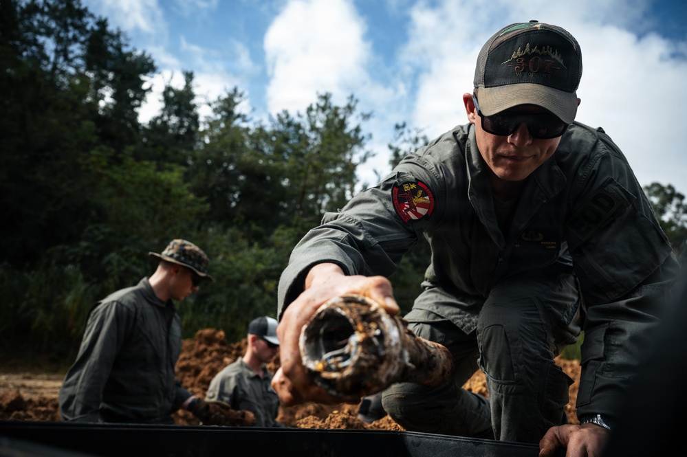Military or EOD (Explosive Ordnance Disposal) personnel are handling what appears to be an excavated ordnance or munition item in an outdoor field setting. The primary figure in the foreground is wearing olive/gray tactical clothing with a unit patch on the sleeve, sunglasses, and a baseball cap, and is holding a corroded cylindrical object. Additional personnel are visible in the background working in an excavated dirt area surrounded by trees.