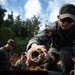 Military or EOD (Explosive Ordnance Disposal) personnel are handling what appears to be an excavated ordnance or munition item in an outdoor field setting. The primary figure in the foreground is wearing olive/gray tactical clothing with a unit patch on the sleeve, sunglasses, and a baseball cap, and is holding a corroded cylindrical object. Additional personnel are visible in the background working in an excavated dirt area surrounded by trees.