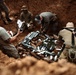 Military personnel are working inside a large excavated pit in red clay soil, handling what appears to be a collection of unexploded ordnance (UXO) or old munitions. The team appears to be conducting an Explosive Ordnance Disposal (EOD) operation, arranging demolition materials including what appear to be detonating cord or explosive strips across the ordnance pile. At least four personnel are visible in and around the pit, working in a coordinated manner.