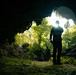 A dramatic silhouette photograph taken from inside a cave looking outward toward a lush, sunlit jungle entrance. A tall figure stands in the foreground holding what appears to be a walking stick or pole, while at least two other individuals are visible crouching or sitting near the cave opening. Strong backlighting from the sun creates a high-contrast composition with vivid green tropical vegetation framing the cave mouth.