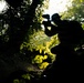 A silhouetted photographer crouches in a dense tropical forest, capturing images with a camera equipped with a lens or flash attachment. The scene is backlit, creating a dramatic contrast between the dark foreground figures and the bright, sunlit green foliage in the background. A second silhouetted figure is partially visible on the left side of the frame.