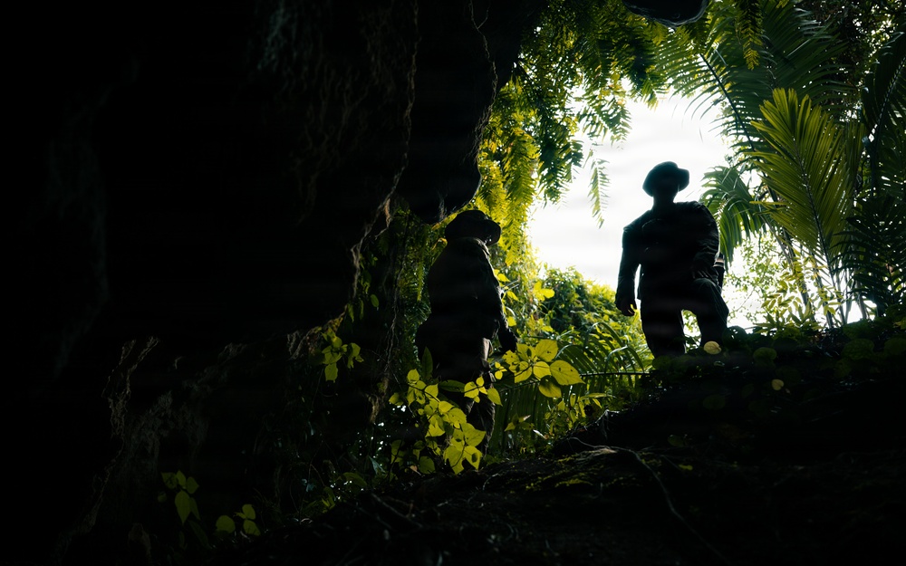 The image is taken from a low angle inside or at the base of a cave or rocky overhang, looking upward toward two silhouetted figures against a bright, overcast sky. The figures appear to be exploring or hiking through a dense tropical jungle environment. The dramatic backlighting creates strong contrast between the dark foreground and the luminous green foliage surrounding the cave opening.