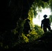 The image is taken from a low angle inside or at the base of a cave or rocky overhang, looking upward toward two silhouetted figures against a bright, overcast sky. The figures appear to be exploring or hiking through a dense tropical jungle environment. The dramatic backlighting creates strong contrast between the dark foreground and the luminous green foliage surrounding the cave opening.