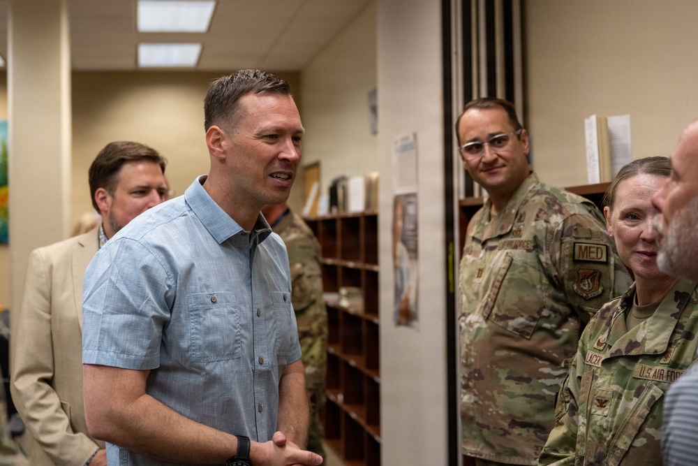A group of individuals, including U.S. Air Force personnel in OCP (Operational Camouflage Pattern) uniforms and civilians in business casual attire, are gathered in what appears to be an office or administrative area on a military installation. A man in a light blue button-down shirt appears to be speaking or presenting to the group. The setting suggests a briefing, tour, or official visit.