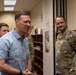A group of individuals, including U.S. Air Force personnel in OCP (Operational Camouflage Pattern) uniforms and civilians in business casual attire, are gathered in what appears to be an office or administrative area on a military installation. A man in a light blue button-down shirt appears to be speaking or presenting to the group. The setting suggests a briefing, tour, or official visit.