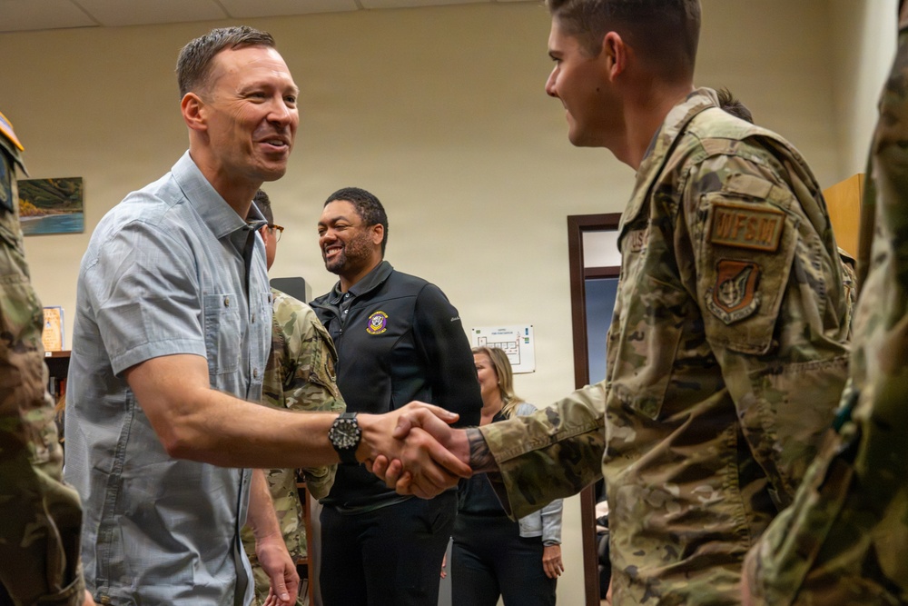 A civilian man in a light blue short-sleeve shirt is shaking hands with a military service member in OCP (Operational Camouflage Pattern) uniform. The setting appears to be an indoor military facility or office environment. Several other individuals, including uniformed personnel and civilians, are visible in the background observing the interaction.