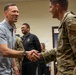 A civilian man in a light blue short-sleeve shirt is shaking hands with a military service member in OCP (Operational Camouflage Pattern) uniform. The setting appears to be an indoor military facility or office environment. Several other individuals, including uniformed personnel and civilians, are visible in the background observing the interaction.