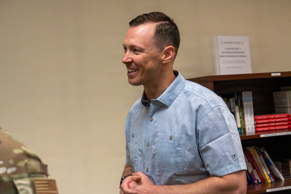 A smiling man in a light blue button-up shirt stands in what appears to be an office or administrative setting. He is engaged in conversation, facing to his left with a broad smile. A bookshelf with various books and an important notice sign is visible in the background, along with a partially visible person in military camouflage uniform.