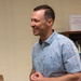 A smiling man in a light blue button-up shirt stands in what appears to be an office or administrative setting. He is engaged in conversation, facing to his left with a broad smile. A bookshelf with various books and an important notice sign is visible in the background, along with a partially visible person in military camouflage uniform.